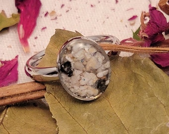 May include: A silver ring with a clear oval-shaped stone filled with white sand and black specks.