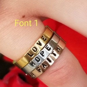 May include: Three stacked rings with the words "LOVE", "HOPE", and "FAITH" engraved on them. The rings are made of different metals, with the top ring being gold, the middle ring being silver, and the bottom ring being a rose gold colour.