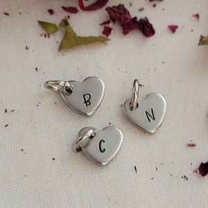 May include: Three small, silver heart-shaped charms with attached loops. Each charm has a single letter: "B", "N", and "C" stamped on the front. The charms are displayed on a white surface with scattered dried flower petals.