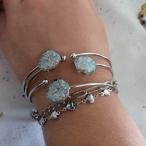 May include: Silver bangle bracelets with iridescent glitter embedded in clear resin circles. The bracelets are stacked on a person's wrist.