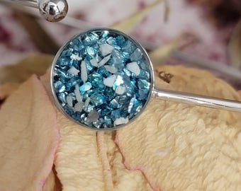 May include: A silver bracelet with a circular charm containing blue and white crushed glass. The charm is set in a silver bezel, and the bracelet has a simple design. The background is a blurred image of dried rose petals. The bracelet is a piece of jewellery.