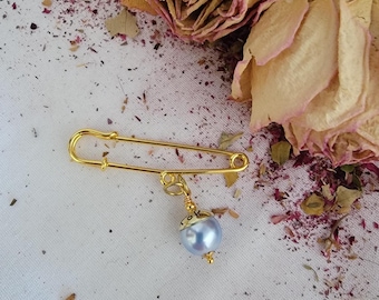 May include: A gold safety pin with a pearl charm hanging from it. The pearl is a light blue color.