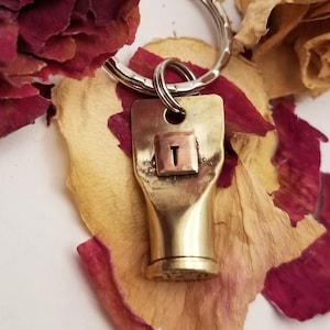 May include: Brass key chain with a letter 'T' charm. The charm is a square with a copper background and a silver letter 'T'. The key chain is on a silver key ring and is surrounded by dried rose petals.