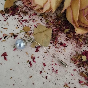 May include: A silver pin with a light blue pearl on a white background with dried rose petals.