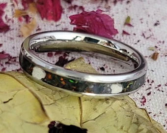 May include: A silver ring with a black and white inlay of crushed opal and other stones. The ring is on a leaf with pink petals in the background.