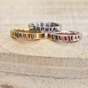 May include: Three personalized rings with the names "Sophia", "Mary", and "Lillian" engraved on them. The rings are stacked on top of each other, with the gold ring on the bottom, the silver ring in the middle, and the rose gold ring on top.