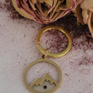 May include: Gold-colored keychain with a circular pendant featuring a mountain range design. The pendant has a cream-colored background and a small black dot. The keychain is set against a backdrop of dried roses.