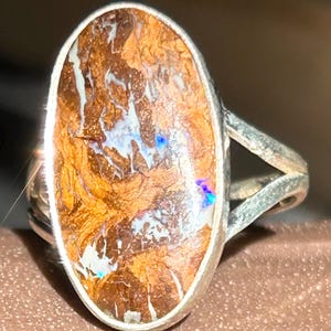 May include: Oval-shaped ring featuring a large, textured opal with a mix of brown, white, and blue hues. The opal is set in a silver-colored band with a split shank design, creating a unique and eye-catching piece of jewelry.