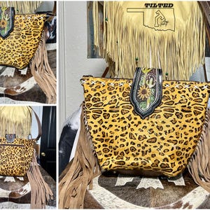 May include: A leopard print handbag featuring long tan fringe on the sides, a brown leather shoulder strap, and a central decorative flap with a sunflower and cactus design. The bag includes white cross-stitching along the top edge.
