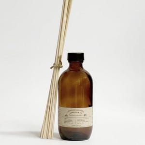 May include: A brown glass aromatherapy diffuser bottle with a black cap and a bundle of wooden reeds tied with twine. The bottle has a label with text. The diffuser is designed for home fragrance.