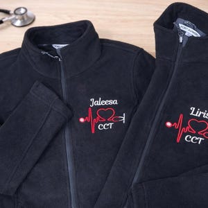 May include: Two black fleece jackets with embroidered medical-themed designs. One jacket has "Jaleesa" and the other "Liris" above a red heartbeat line, heart, and stethoscope graphic. A stethoscope rests in the background.