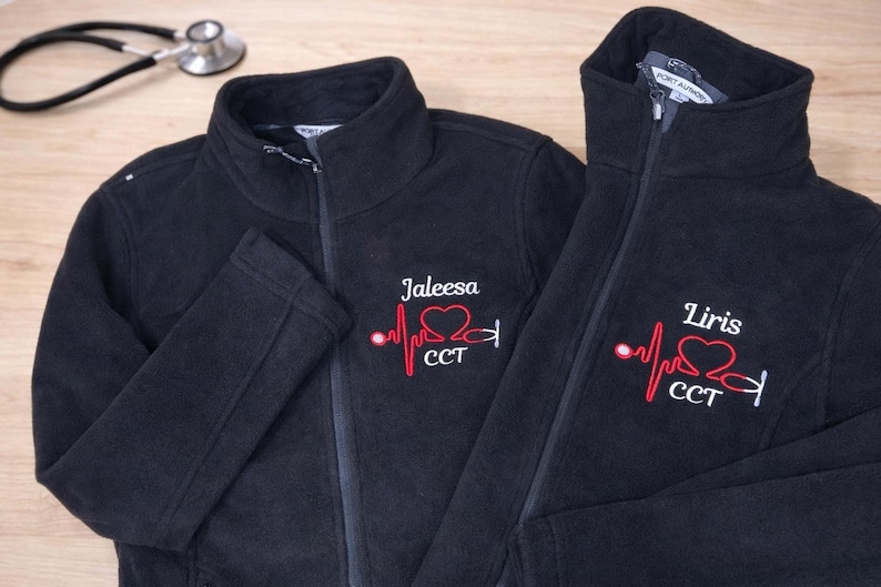 May include: Two black fleece jackets with embroidered medical-themed designs. One jacket has "Jaleesa" and the other "Liris" above a red heartbeat line, heart, and stethoscope graphic. A stethoscope rests in the background.