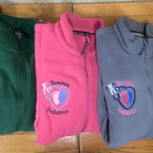 May include: A row of fleece zip-up jackets in black, green, pink, and gray. Each jacket features an embroidered logo with a heart, stethoscope, and the words "Bambini Pediatrics". The jackets are folded and displayed on a wooden surface.