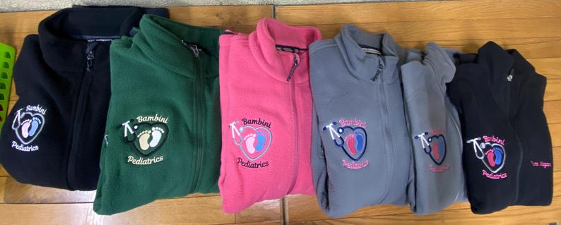 May include: A row of fleece zip-up jackets in black, green, pink, and gray. Each jacket features an embroidered logo with a heart, stethoscope, and the words "Bambini Pediatrics". The jackets are folded and displayed on a wooden surface.