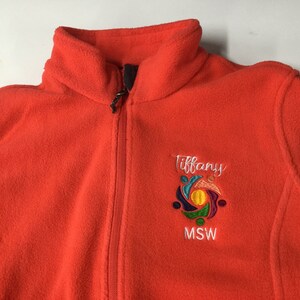 Puede incluir: Una chaqueta de forro polar naranja brillante con cremallera frontal completa y cuello alto. El pecho izquierdo presenta un logotipo bordado con el nombre Tiffany en letra blanca sobre un gr&aacute;fico circular colorido y el texto MSW debajo.