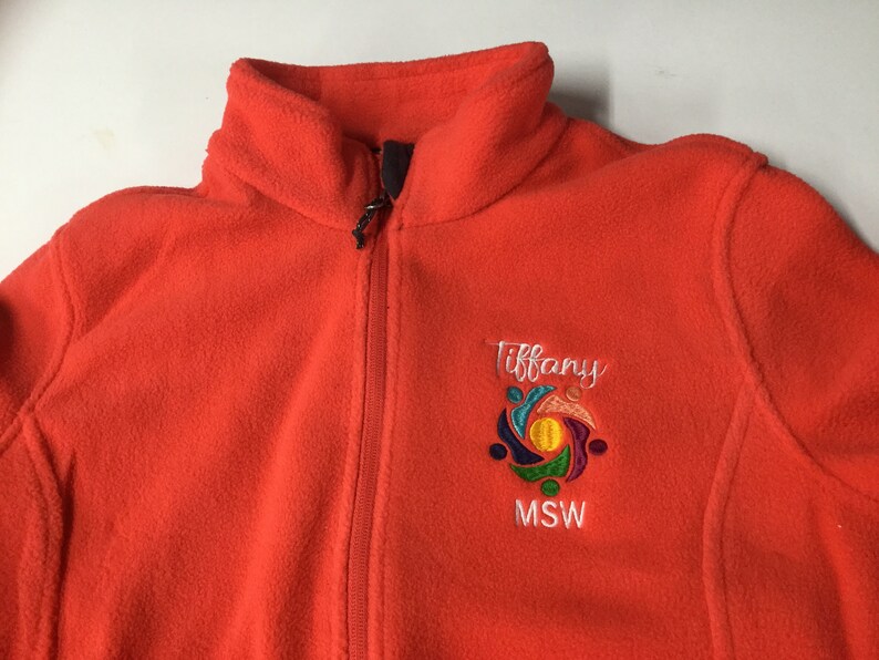Puede incluir: Una chaqueta de forro polar naranja brillante con cremallera frontal completa y cuello alto. El pecho izquierdo presenta un logotipo bordado con el nombre Tiffany en letra blanca sobre un gr&aacute;fico circular colorido y el texto MSW debajo.