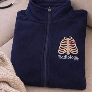 May include: A navy blue fleece zip-up jacket featuring an embroidered beige rib cage graphic on the left chest with a small red heart accent. The word Radiology is embroidered in white script lettering directly below the rib cage design.