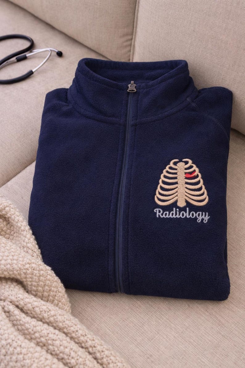 May include: A navy blue fleece zip-up jacket featuring an embroidered beige rib cage graphic on the left chest with a small red heart accent. The word Radiology is embroidered in white script lettering directly below the rib cage design.