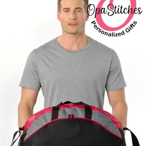 May include: A black and gray duffel bag with red accents. The bag has a zippered main compartment and a front pocket. The bag is being held by a person.