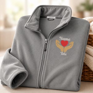 May include: A gray fleece zip-up jacket featuring an embroidered design on the left chest. The embroidery depicts two tan hands cupping a red heart, with the word Name above the heart and the word Title below the heart in white script lettering.
