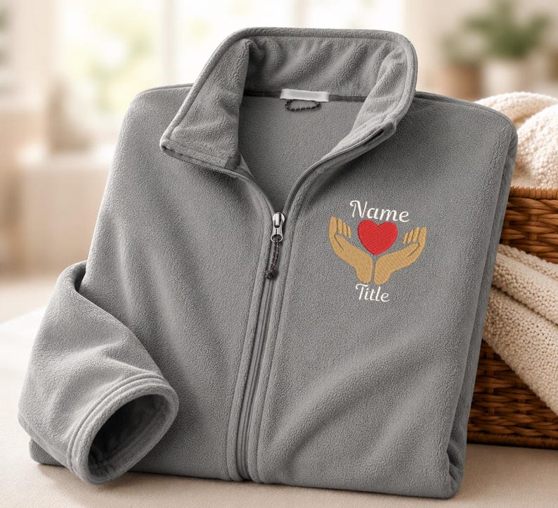 Puede incluir: Una chaqueta de forro polar gris con cremallera y un bordado en el pecho izquierdo. El dise&ntilde;o muestra dos manos de color beige sosteniendo un coraz&oacute;n rojo, con la palabra Name sobre el coraz&oacute;n y la palabra Title debajo en letra cursiva blanca.