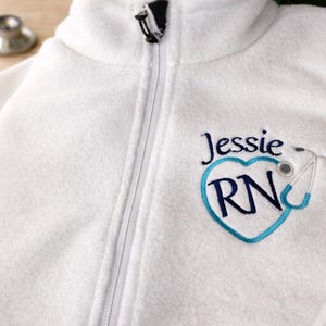 May include: White fleece zip-up jacket with embroidered design. The design includes the name "Jessie", a blue heart with "RN" inside, and a stethoscope. A stethoscope is visible in the background.