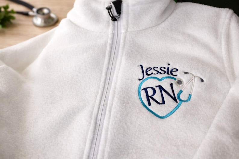 May include: White fleece zip-up jacket with embroidered design. The design includes the name "Jessie", a blue heart with "RN" inside, and a stethoscope. A stethoscope is visible in the background.