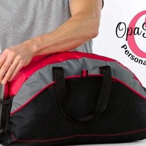 May include: A person is putting a silver water bottle into the side pocket of a black, red, and gray duffel bag. The bag has a red trim and two black handles. The bag is sitting on a white surface. The text " Opa Stitches Personalized Gifts" is in the upper right corner of the image.