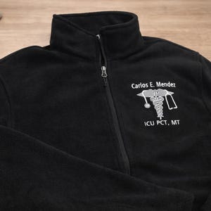 May include: Black fleece zip-up jacket with a high collar. The jacket features a white embroidered medical symbol and the text "Carlos E. Mendez" and "ICU PCT, MT".