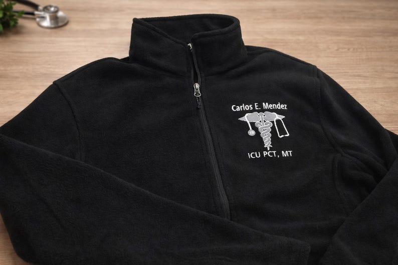 May include: Black fleece zip-up jacket with a high collar. The jacket features a white embroidered medical symbol and the text "Carlos E. Mendez" and "ICU PCT, MT".
