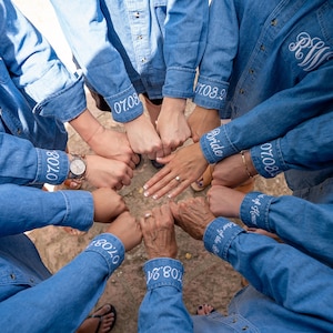 May include: A group of people wearing blue denim shirts with white embroidery. The shirts have the date "07.08.2019" and the word "Bride" embroidered on the cuffs. The people are holding hands in a circle.