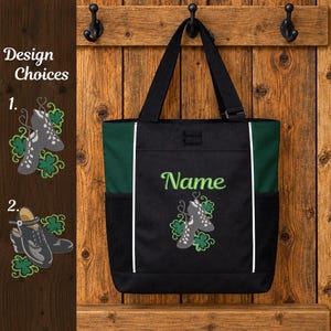 May include: A black and dark green tote bag featuring an embroidered design of two grey dance shoes surrounded by green shamrocks. The word Name is embroidered in light green above the shoes. The bag has black mesh side pockets.