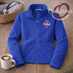 May include: Royal blue fleece jacket with a full zipper, two side pockets, and a stand-up collar. The jacket has an embroidered design of a heartbeat and a cross, with the words "Name" and "Title" above and below the design. A white mug with a heart-shaped design is on the left.