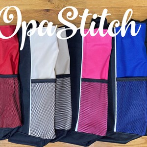 May include: A row of black tote bags with colored side panels and mesh pockets. The side panels are red, beige, gray, pink, blue, and green. Each bag has a white stripe and the text "Opa Stitch" in a cursive font.