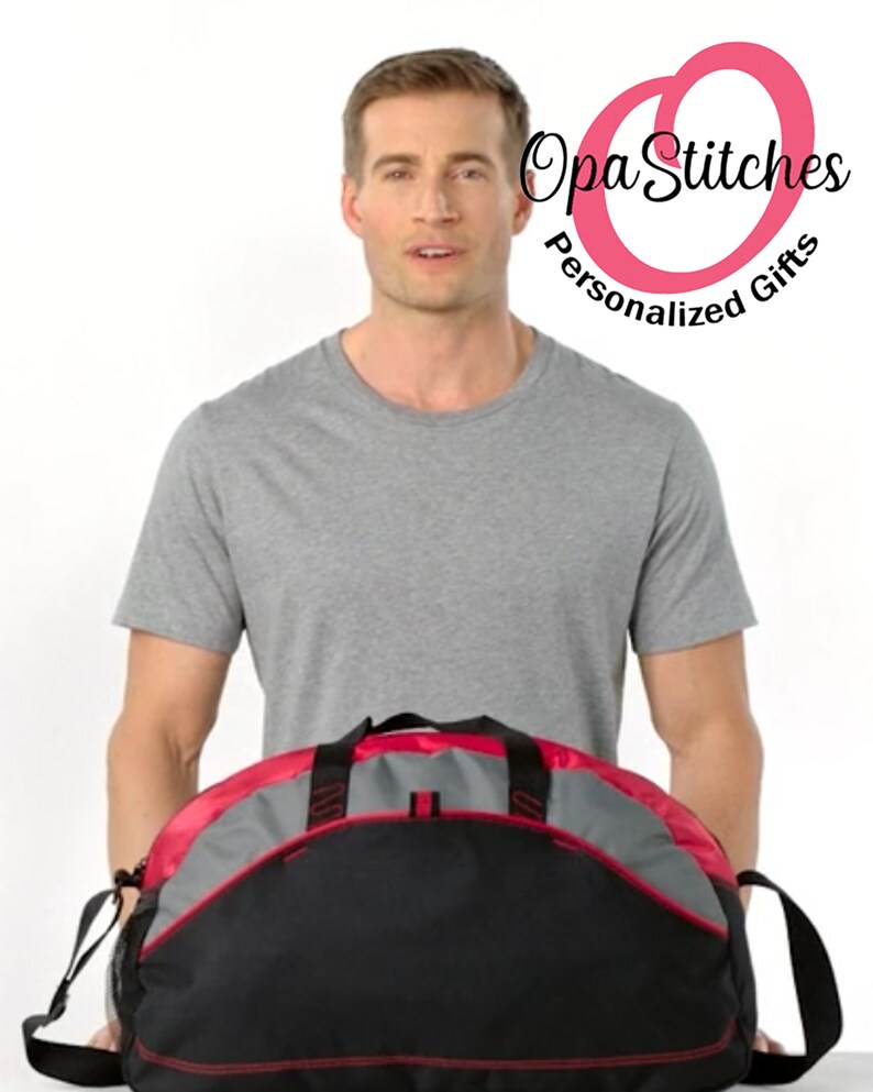 May include: A black and grey duffel bag with red accents. The bag has a zipped main compartment and a front pocket. The bag is being held by a person.