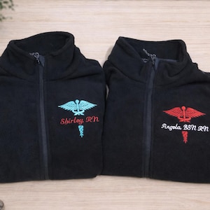 May include: Two black fleece jackets with a zipper closure. Each jacket has an embroidered medical symbol and a name with credentials. One has a teal symbol and the name "Shirley, RN". The other has a red symbol and the name "Angela, BSN, RN".