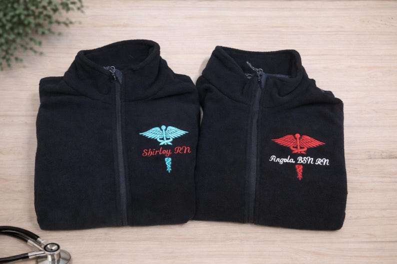 May include: Two black fleece jackets with a zipper closure. Each jacket has an embroidered medical symbol and a name with credentials. One has a teal symbol and the name "Shirley, RN". The other has a red symbol and the name "Angela, BSN, RN".