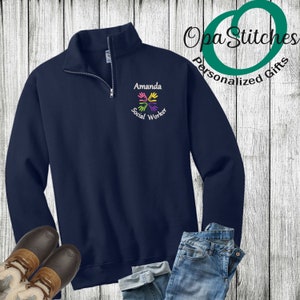 May include: Navy blue quarter-zip sweatshirt with the name "Amanda" and "Social Worker" embroidered in white. A colorful handprint design is embroidered below the name. The sweatshirt is paired with blue jeans and brown boots.