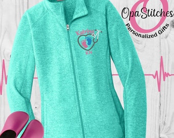 Könnte beinhalten: Eine türkisblaue Fleecejacke mit Reißverschluss. Die Jacke hat ein pinkfarbenes Herz mit einem Stethoskop und gestickten Babyfüßen auf der linken Brust. Der Text "Katrina RN" ist unter dem Herzen gestickt.