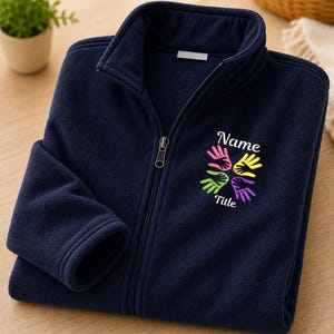 Puede incluir: Una chaqueta polar azul marino con cremallera, cuello alto y cierre frontal completo. En el pecho lleva bordado el texto "Name" y "Title" rodeando un gr&aacute;fico colorido de cuatro huellas de manos en colores rosa, amarillo, verde y morado.