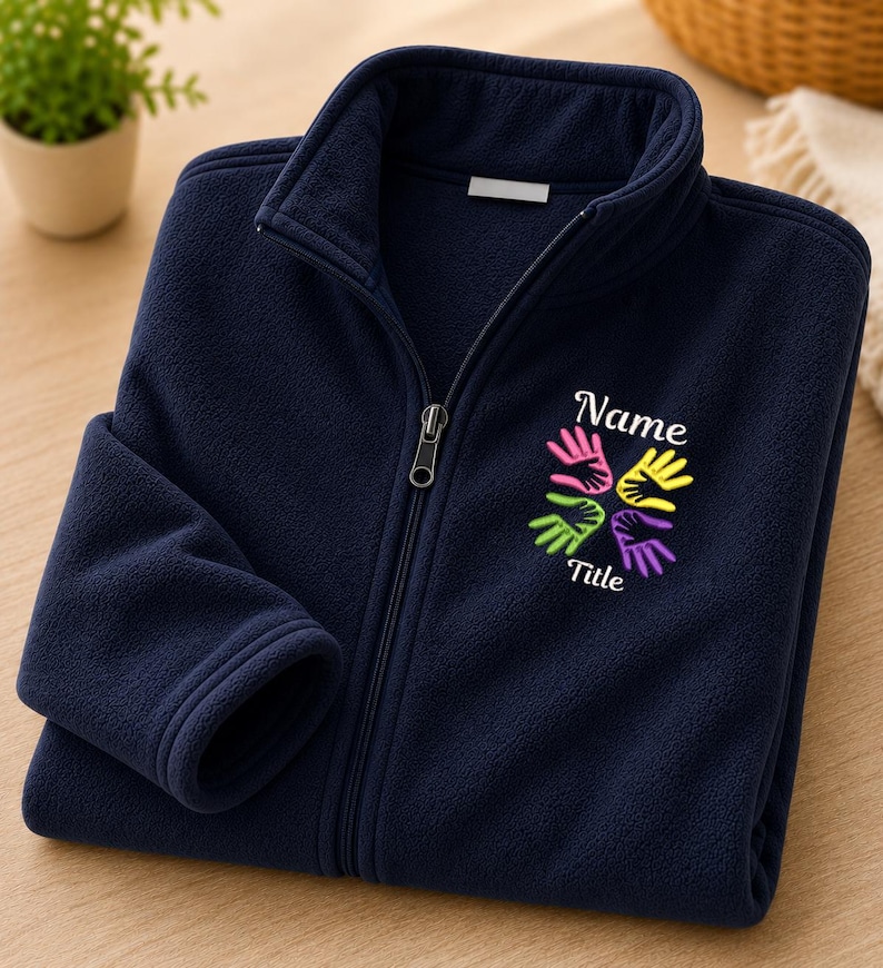 Puede incluir: Una chaqueta polar azul marino con cremallera, cuello alto y cierre frontal completo. En el pecho lleva bordado el texto "Name" y "Title" rodeando un gr&aacute;fico colorido de cuatro huellas de manos en colores rosa, amarillo, verde y morado.