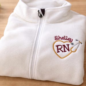 May include: White fleece zip-up jacket with a high collar. The jacket features an embroidered heart design with the name "Shelley", the letters "RN", and a stethoscope. The jacket is folded on a wooden surface.