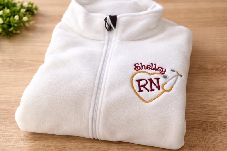 May include: White fleece zip-up jacket with a high collar. The jacket features an embroidered heart design with the name "Shelley", the letters "RN", and a stethoscope. The jacket is folded on a wooden surface.