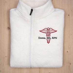 May include: A folded white fleece zip-up jacket with a black zipper. The jacket features a red embroidered caduceus symbol and the text "Emma, MS, RPN". A stethoscope rests in the background.
