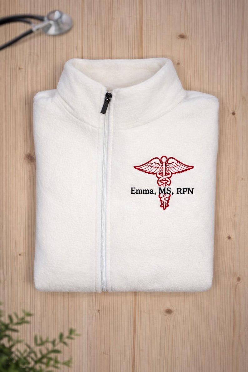 May include: A folded white fleece zip-up jacket with a black zipper. The jacket features a red embroidered caduceus symbol and the text "Emma, MS, RPN". A stethoscope rests in the background.