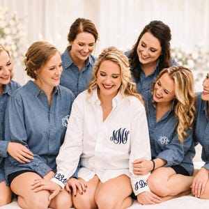 May include: A group of seven individuals wearing long-sleeved button-down shirts. One person wears a white shirt with navy blue monogrammed embroidery on the pocket and cuff. Six others wear blue denim shirts with white monogrammed embroidery.