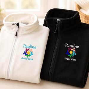 Puede incluir: Dos chaquetas de forro polar, una blanca y otra negra, con cremallera frontal completa y cuello alto. Cada chaqueta muestra un logotipo bordado con el nombre Pauline y el texto Social Work sobre un icono circular de figuras humanas.