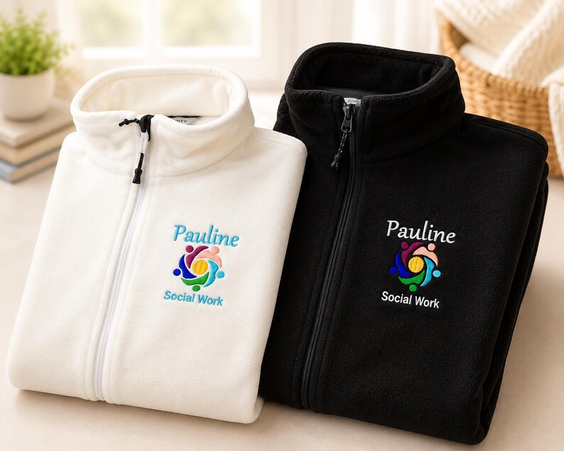 Puede incluir: Dos chaquetas de forro polar, una blanca y otra negra, con cremallera frontal completa y cuello alto. Cada chaqueta muestra un logotipo bordado con el nombre Pauline y el texto Social Work sobre un icono circular de figuras humanas.