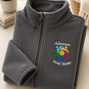 Puede incluir: Una chaqueta de forro polar gris oscuro con cremallera frontal completa y cuello alto. El pecho presenta un logotipo bordado de figuras coloridas interconectadas que rodean un c&iacute;rculo amarillo, con el texto Adrienne Social Worker debajo.