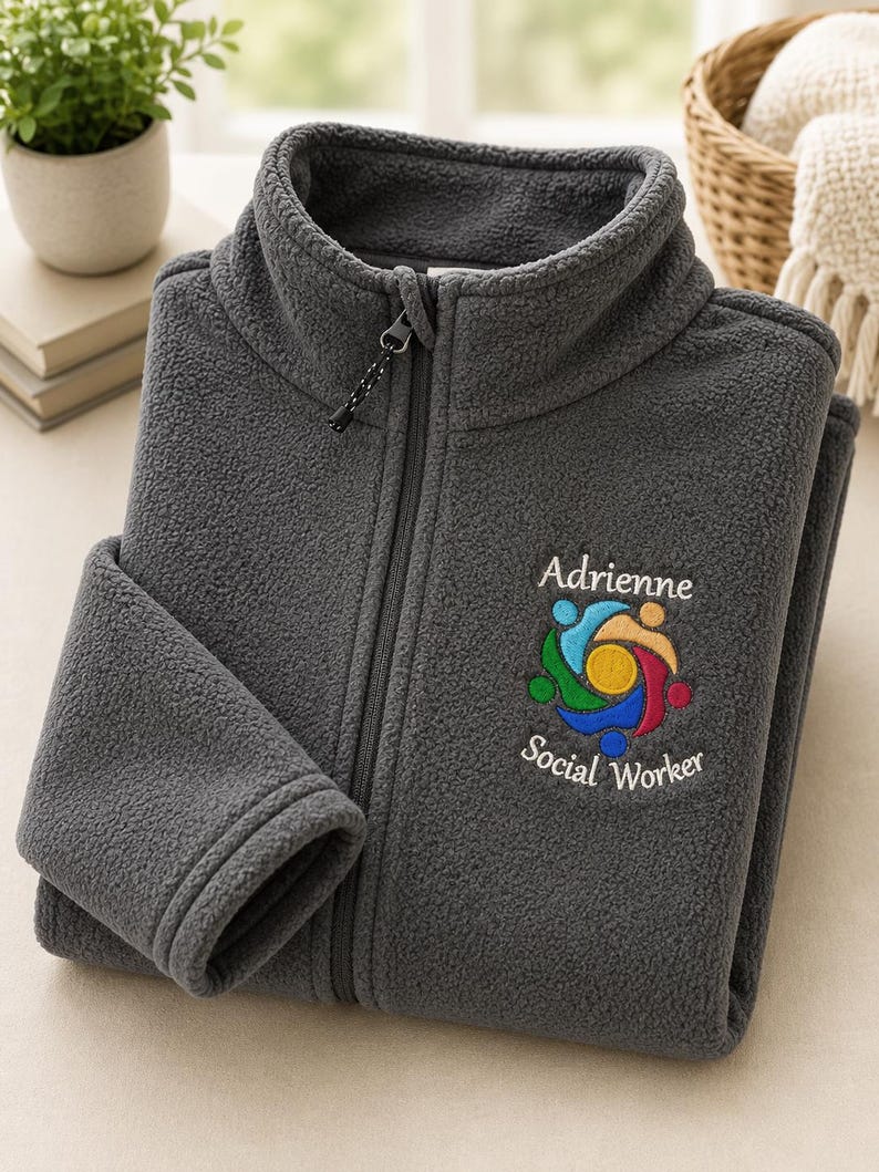 Puede incluir: Una chaqueta de forro polar gris oscuro con cremallera frontal completa y cuello alto. El pecho presenta un logotipo bordado de figuras coloridas interconectadas que rodean un c&iacute;rculo amarillo, con el texto Adrienne Social Worker debajo.